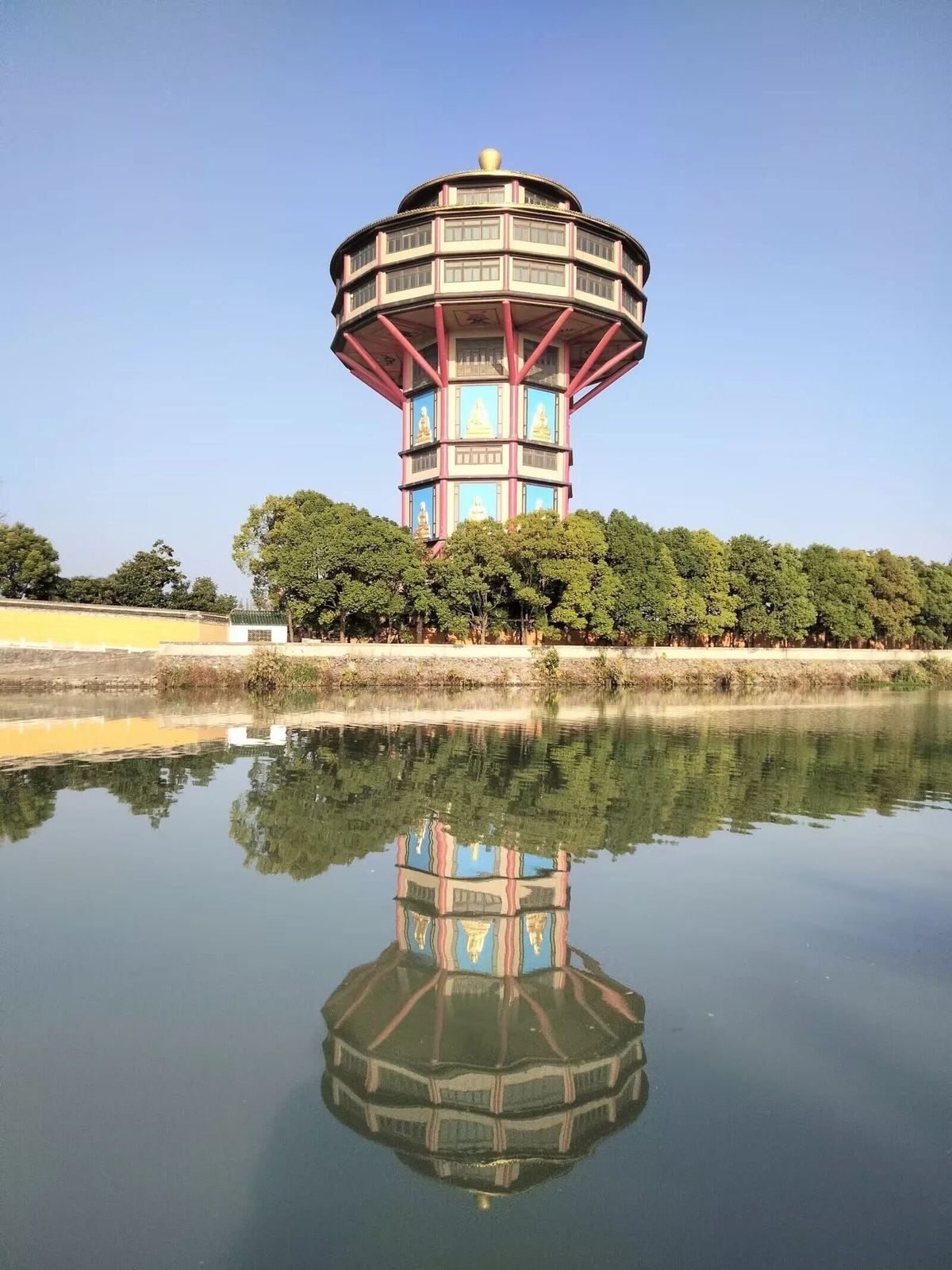 Torre circular de faixas vermelhas e painéis azuis refletida na água parada do canal